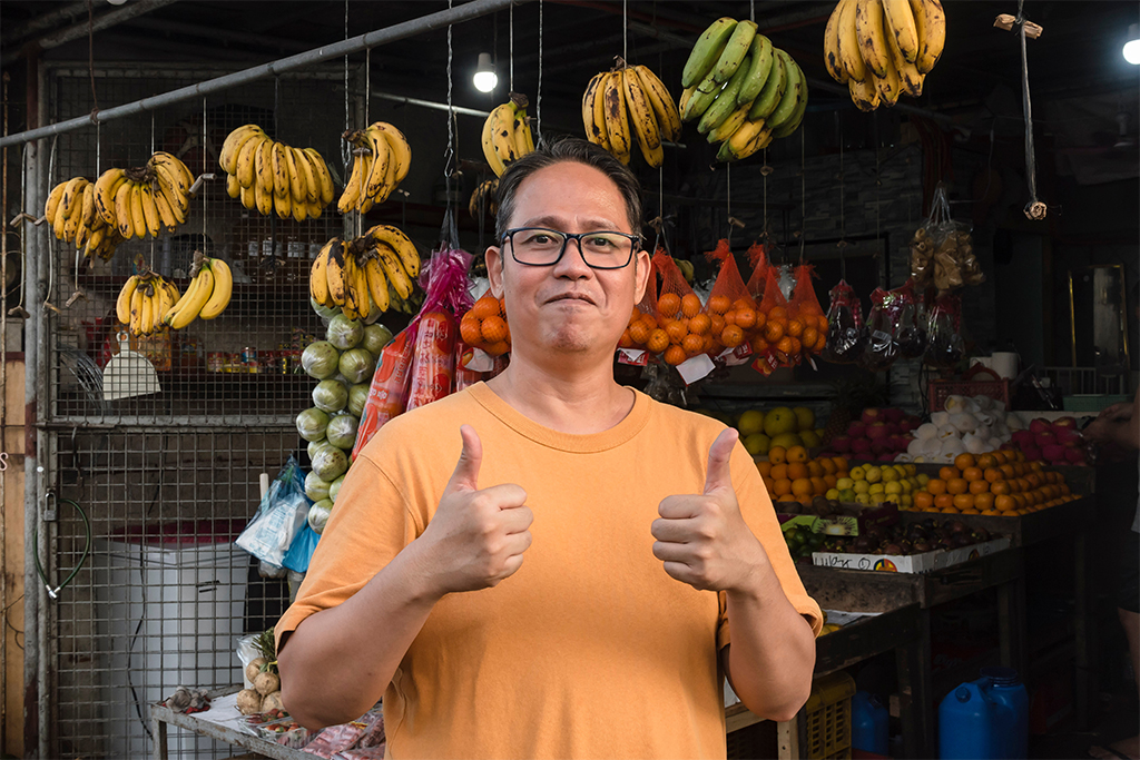 A middle aged Filipino fruit vendor, posing in front of his open-air store, making a thumbs up sign. A small business owner