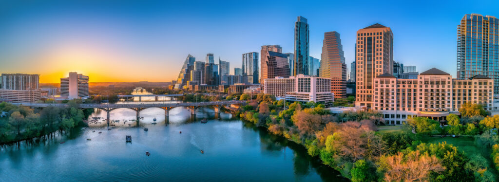 Austin, Texas- Panoramic cityscape and Colorado River against the sunset sky. There are bridges over the river with boats under and a view of the skyscrapers on the sides.